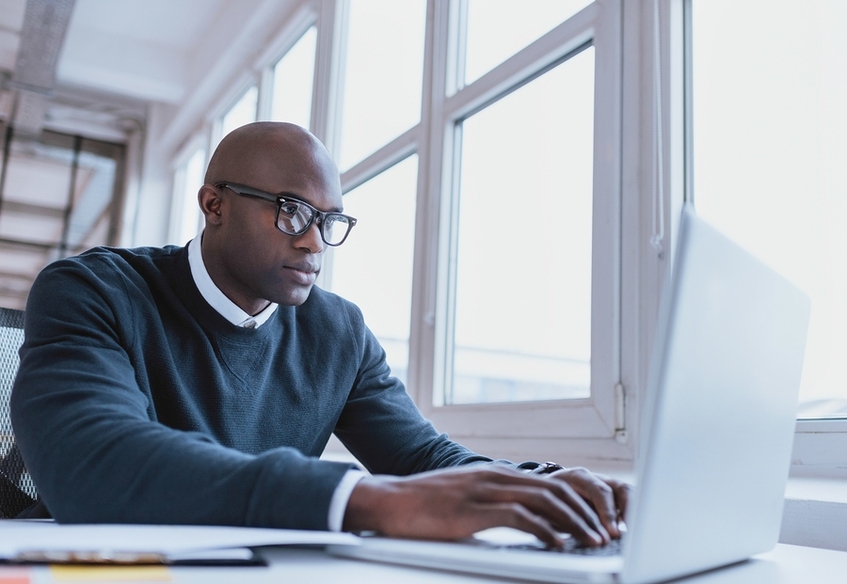 Man with blue sweater over white collar shirt typing on laptop.